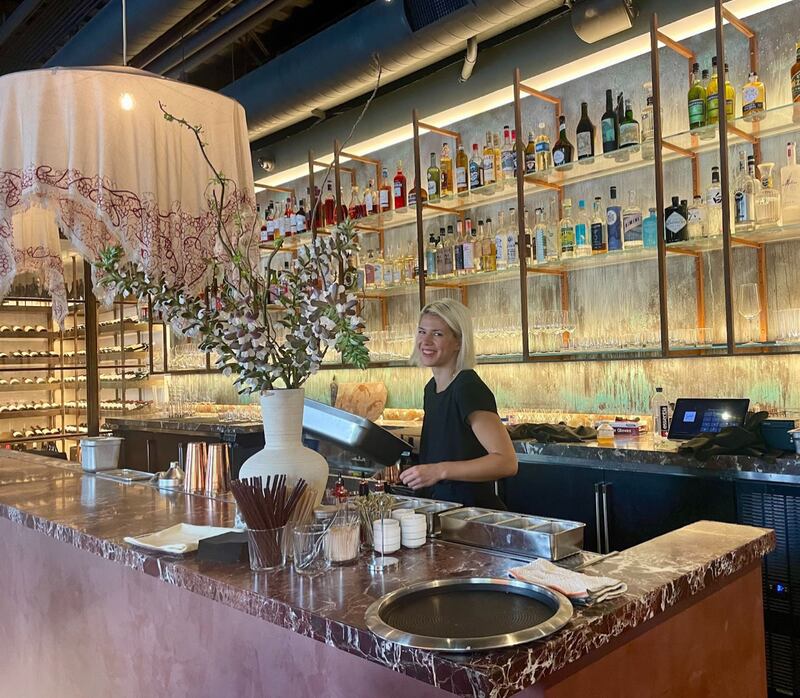 The woman behind the bar smiles at the camera. Liquor bottle are on gold shelves behind her and the bar is a made of black marble.