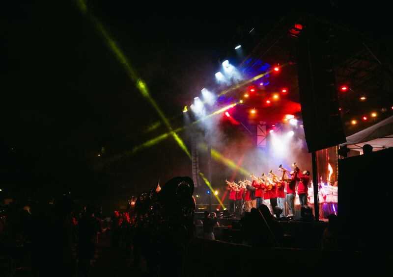 The band is wearing red blazers and at the front of the stage. It is dark out but there are listeners in the crowd while lights shine on stage
