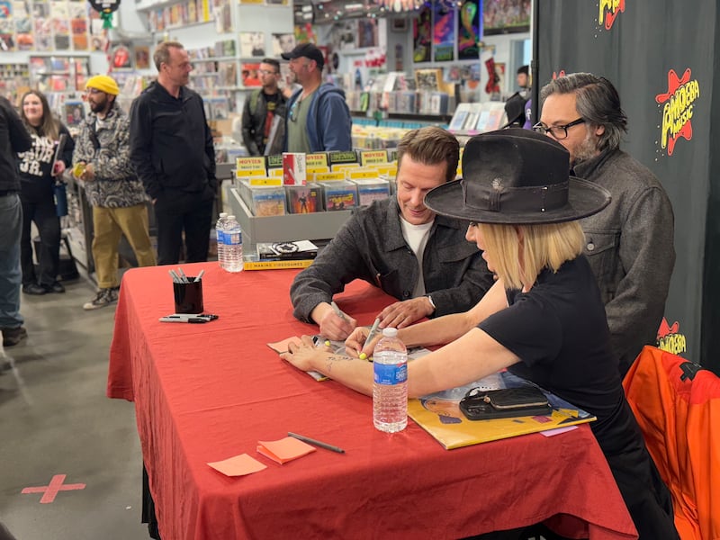 Photo of two people signing an album at a table with a person standing behind them