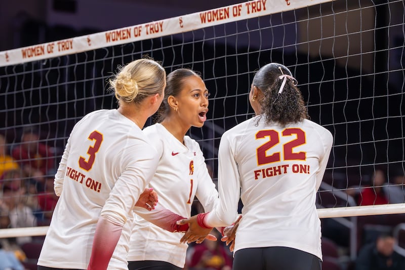 Faumuina (1) celebrates a point with Reese Messer (3) and Rylie McGinest (22) behind the net. All wear white USC volleyball uniforms with cardinal and gold accents.