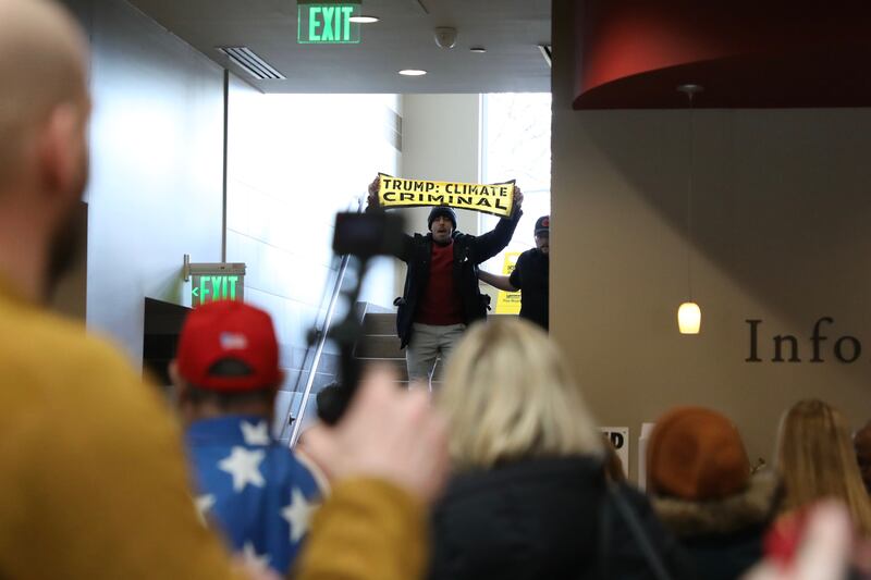 A crowd stands with their backs to the camera in the foreground. They are indoors and a staircase is visible. A man, walking down the stairs, holds a yellow sign reading "Trump: Climate Criminal."