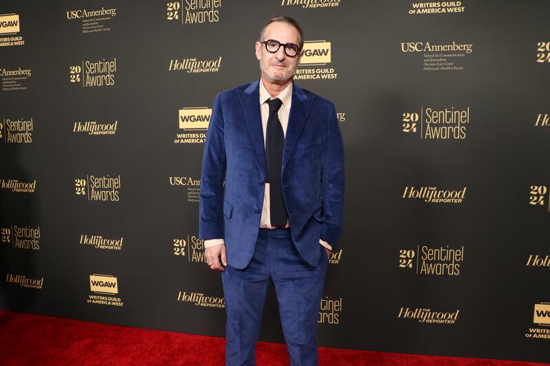 John is weaing a navy blue, felt suit. He has a black tie, cream undershirt and glasses.