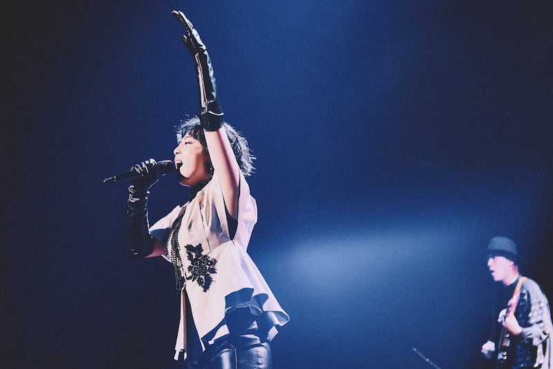 Laco, in a white structured blouse and black pants, holds the mic in one hand and raises the other to the audience.