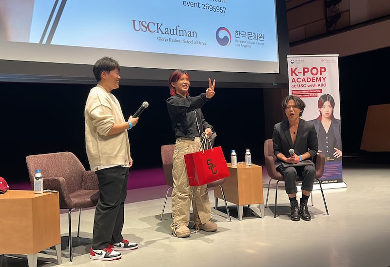 Photo of three people on stage with one signing a v