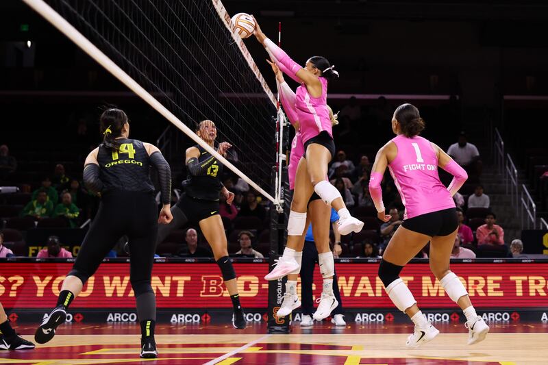 Two USC volleyball players in pink and black uniforms jump up to block a ball over the next against two Oregon players.