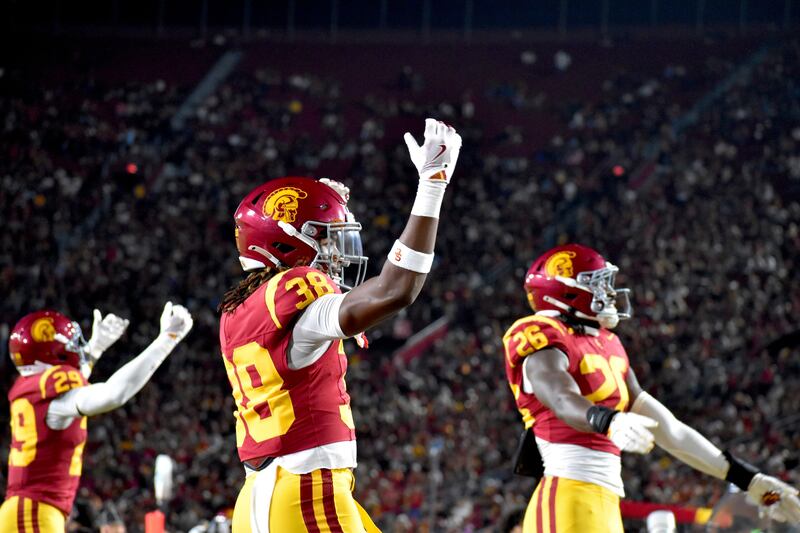 USC football players stand on field with arms outstretched to amp up the Coliseum crowd. They wear cardinal and gold football uniforms.