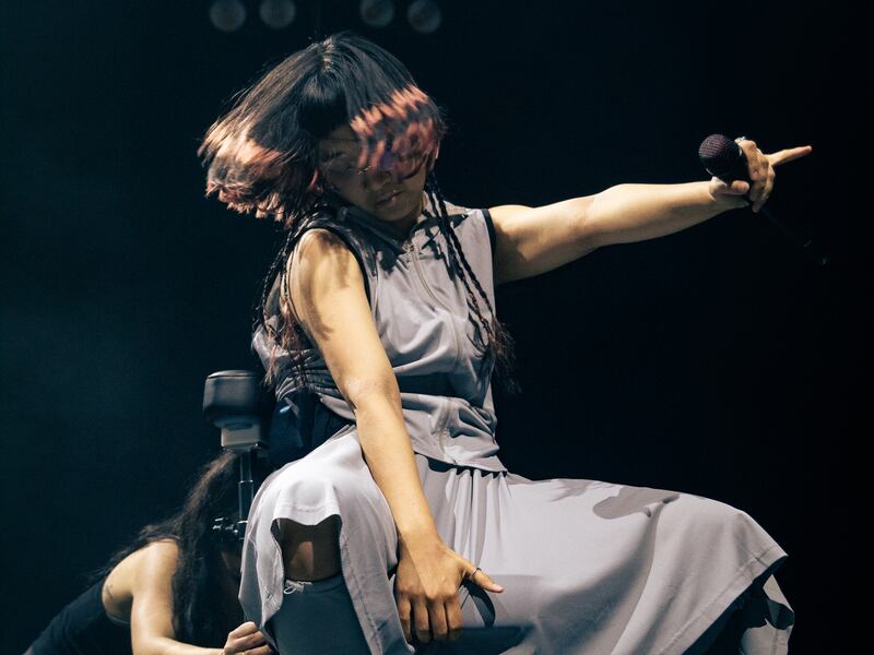 Photo of a woman on a rolling chair with a microphone. Another person is pushing the chair behind her
