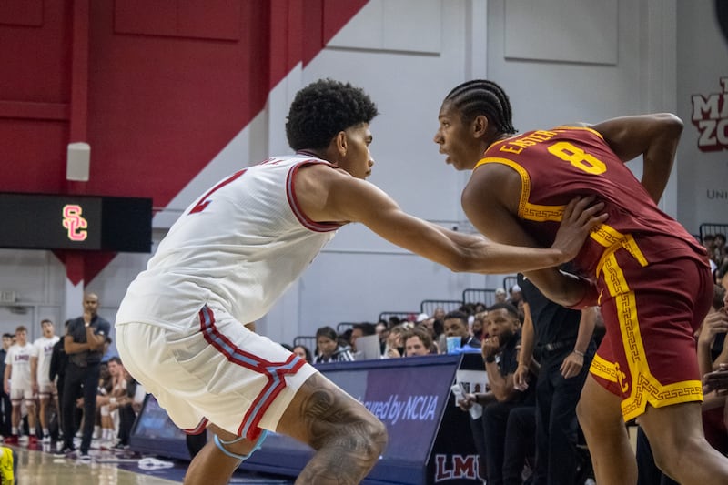 Jerry Easter II (8) faces off with an LMU defender; easter is poised with the ball in his hands off to the side. He wears a cardinal and gold basketball uniform, while the defender wears a white basketball uniform with blue and red accents.