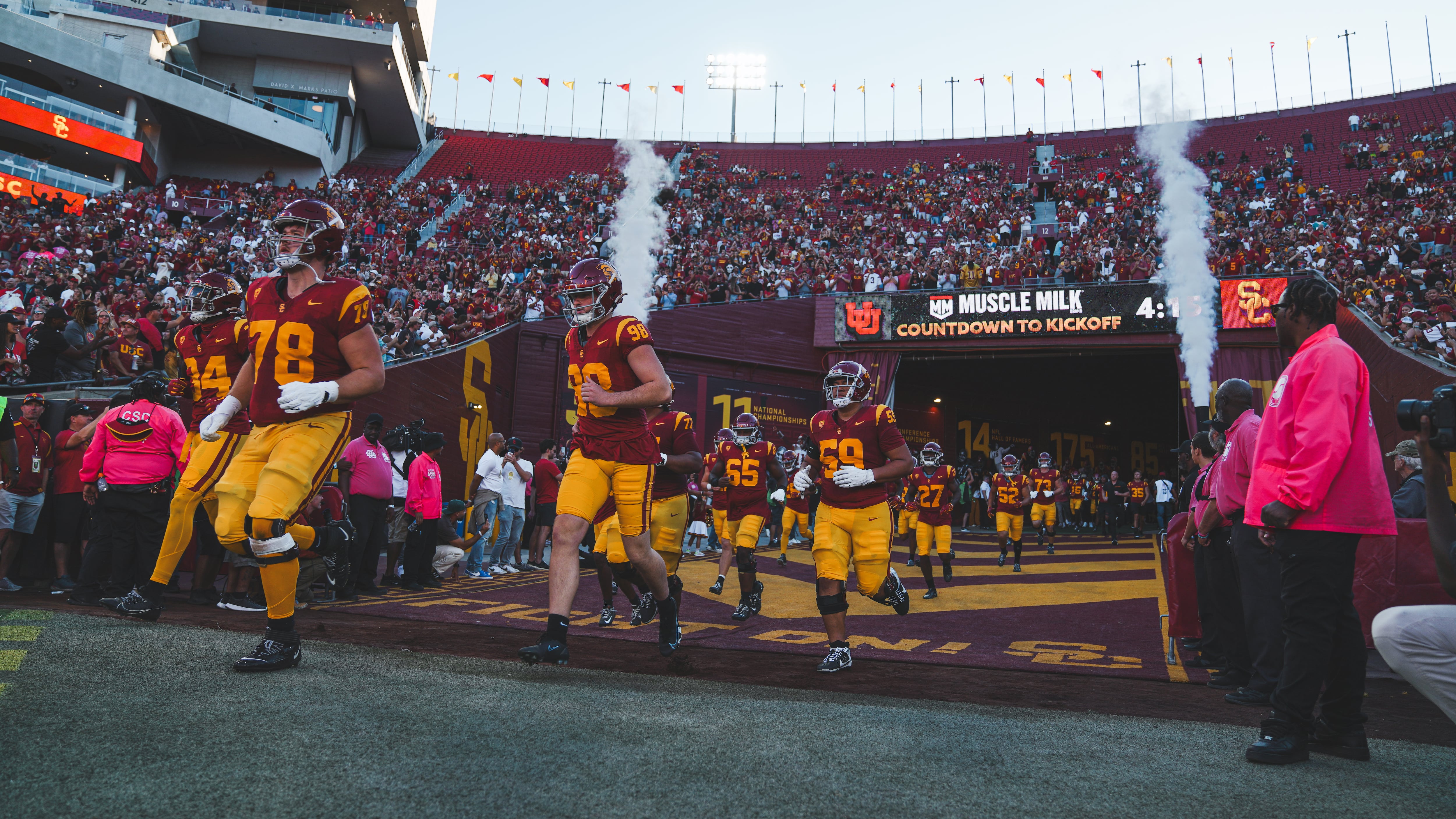 USC's football team runs out onto the field, led by No. 78 Andrew Milek and No. 98 Garth White. They and the rest of the team are framed by two plumes of smoke. The stands behind them are full to a point, but the last eight or so rows are empty.