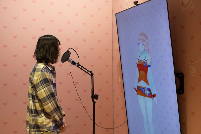 Photo of a person in a flannel shirt speaking into a microphone in front of a screen with an anime girl on it