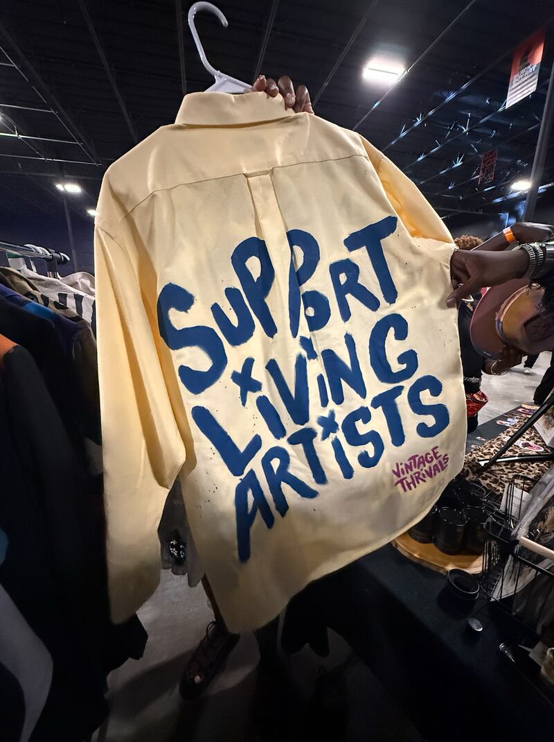 A cream colored jacket hangs on a rack inside an indoor event space. Large blue hand painted letters on the back read “Support Living Artists,” with smaller text in the corner that says “Vintage Thrivals.”