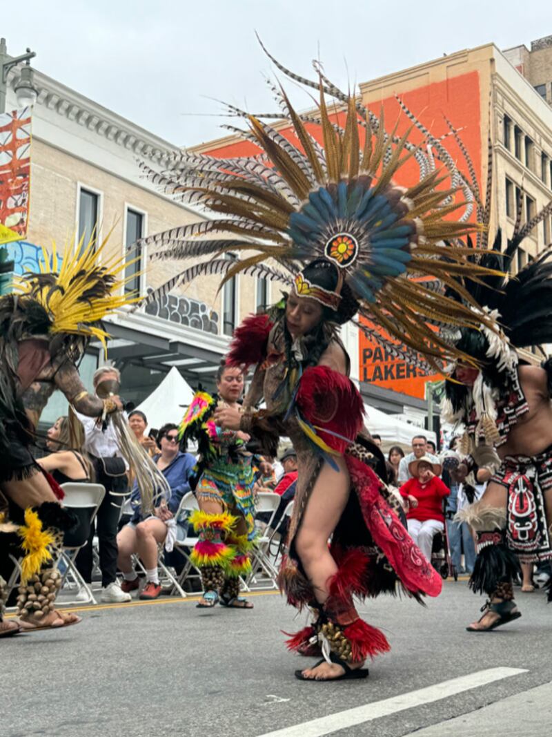 A woman in a red feather dress and a blue and yellow large feather headpiece dances in the street alongside other members of the parade