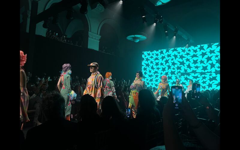 Models in colorful outfits walk down a runway with a blue backdrop with black stars.
