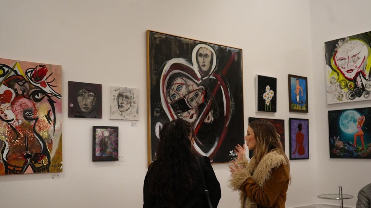 The white walls are covered in different-sized paintings. Two women in coats talk while facing the art.