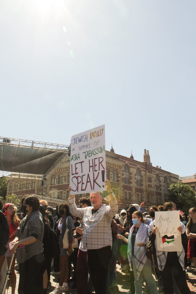 A man in a patterned buttoned down shirt holds up a white sign that says "Jewish Faculty in support of Asna Tabassum" followed by "LET HER SPEAK!" He is surrounded by other protestors as they march along a brick pathway.