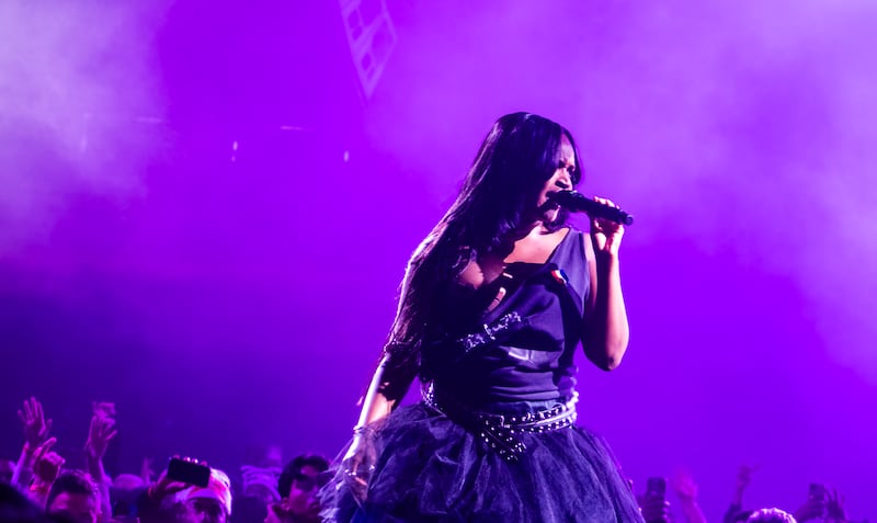 Photo of Amaarae singing with an off-the-shoulder dress. The lights are purple behind her and the crowd has their hands up and is watching her.