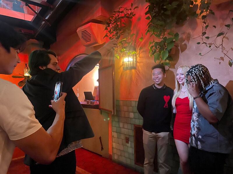 Huey wears a black sweater with a red heart and the letter A in the corner. A blonde woman wears a red dress and the man next to her wears a gray top. They are being photographed against a wall with faux leaves.