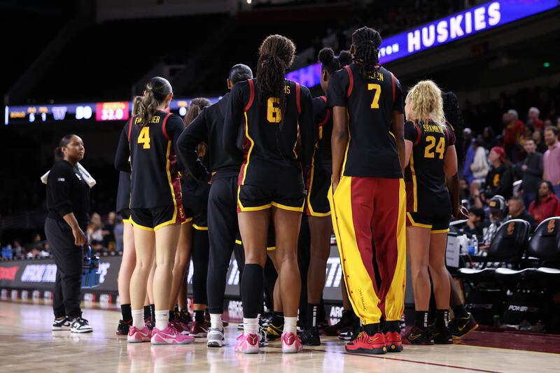 A USC huddle on the sideline. The players wear black basketball uniforms with cardinal and gold accents.