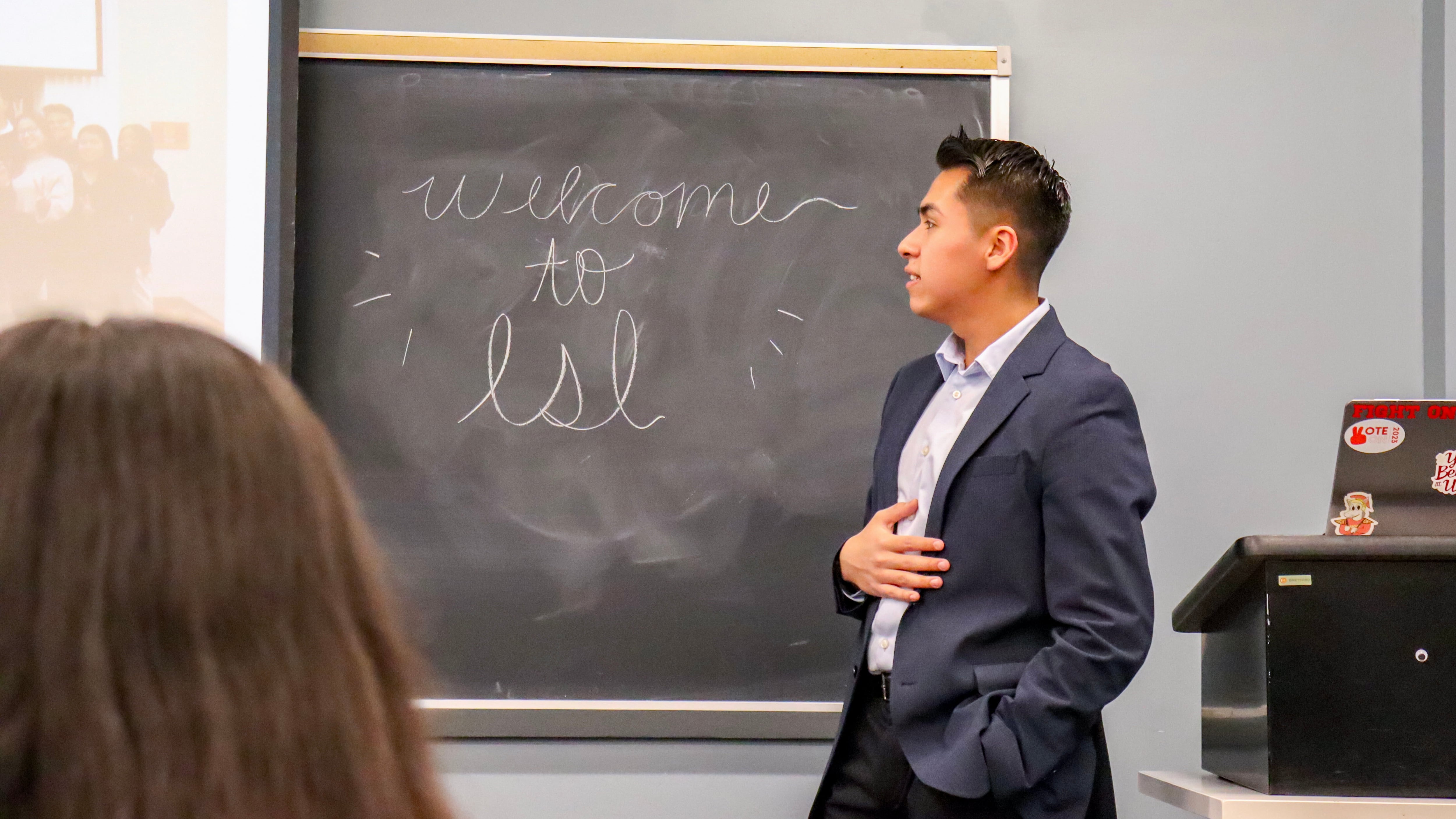 Zuniga wears a dark blue suit, looks at the horizon, there's a blackboard with "welcome to LSL" written on it.