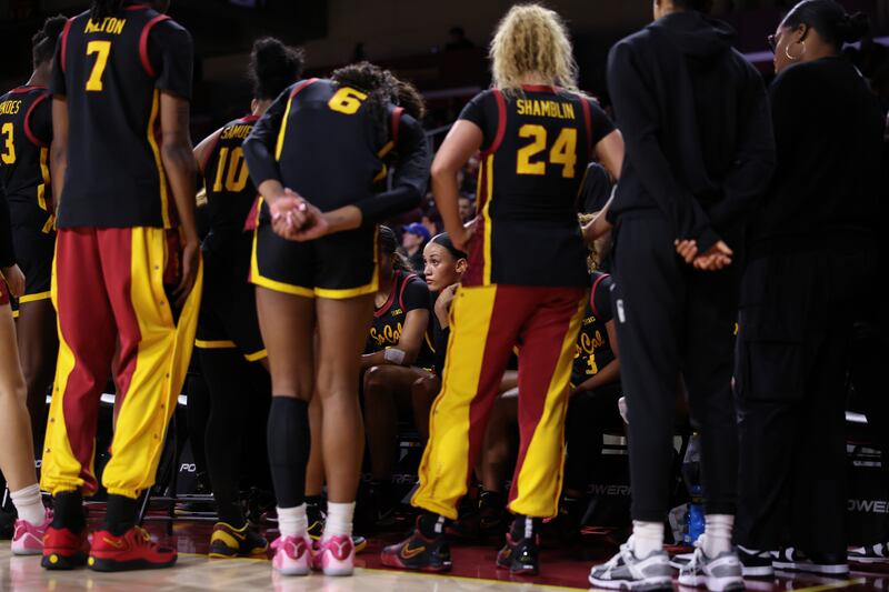 USC players in a sideline huddle wearing black basketball uniforms with cardinal and gold accents.