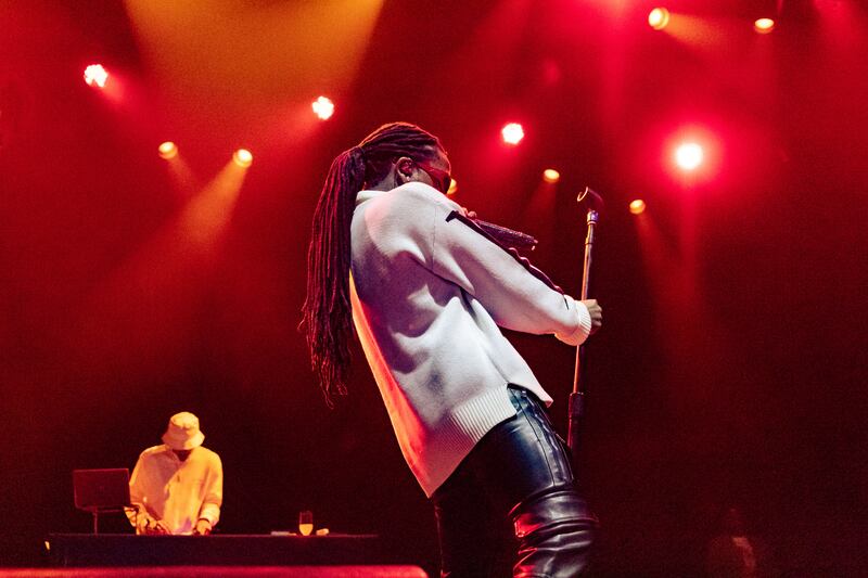A photo of Jacquees holding onto the microphone stand and moving his lower body towards the stand. He is holding the microphone in his other hand and singing into it.