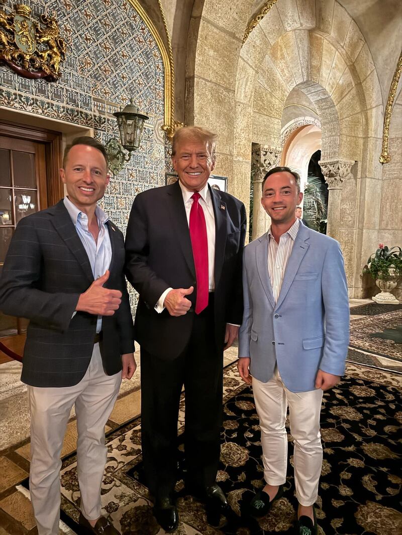 Two men in suits pose with a smiling Donald Trump, who gives the camera a thumbs up.