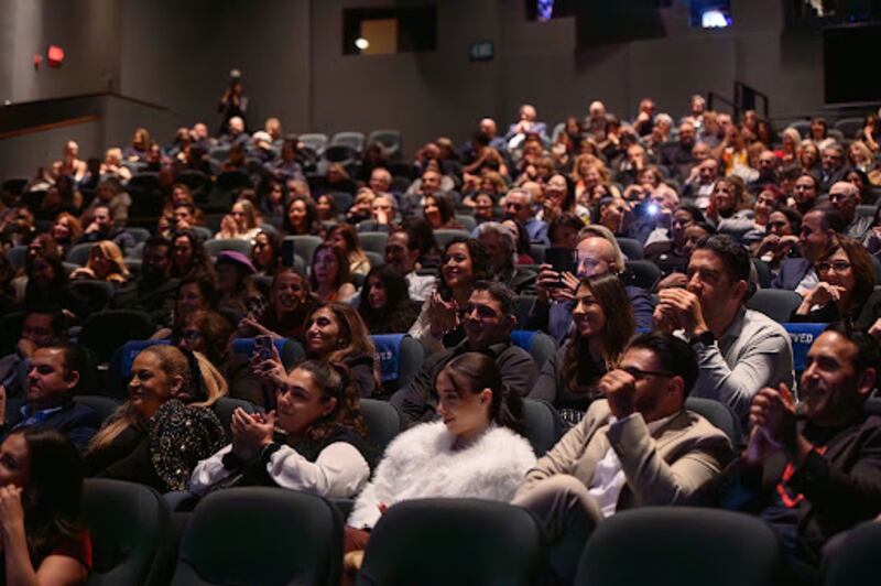 Audience clapping and smiling in dimly lit theater, watching a big screen.