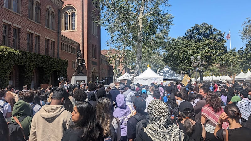 A large group of people gather near a metal statue of a Trojan, white tents are set up in the background for a different event, adding to the feeling of a packed environment.