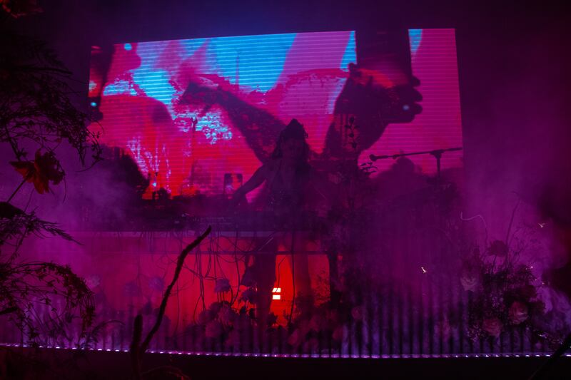 A photo of Arca at her DJ station with pink and blue lights behind her on the projection at the back of the stage.