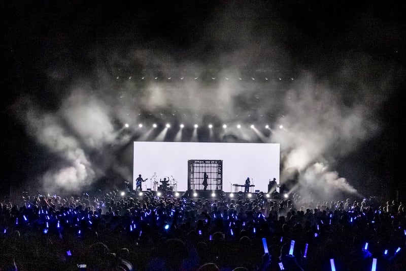 Photo of a crowd of people holding blue sticks in front of a stage with a cage-like cube and a silhouette of a woman in front of the screen