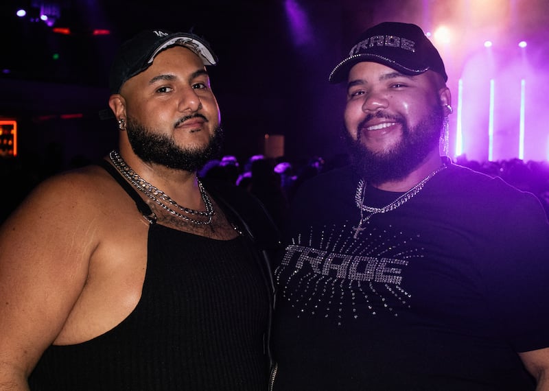 Photo of two concertgoers smiling at the camera. They are both wearing black caps and silver jewelry. The person on the right is wearing a bejeweled shirt that says "Trade" and the person on the left is wearing a blank tank top.