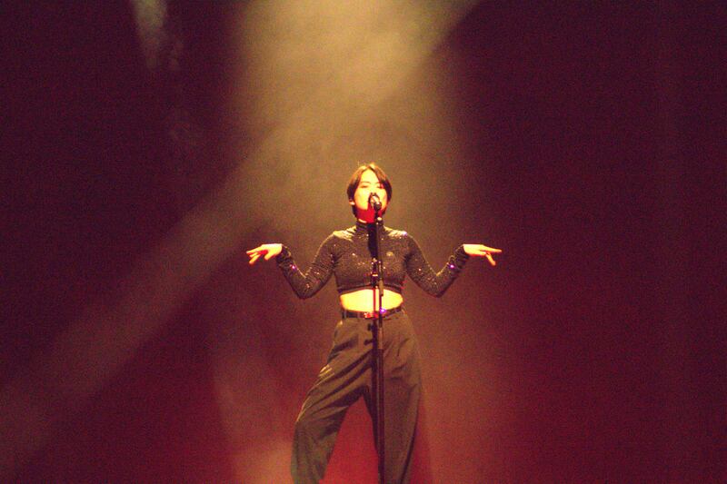 Photo of a woman singing under a spotlight on a red stage with her arms out and wrists bent