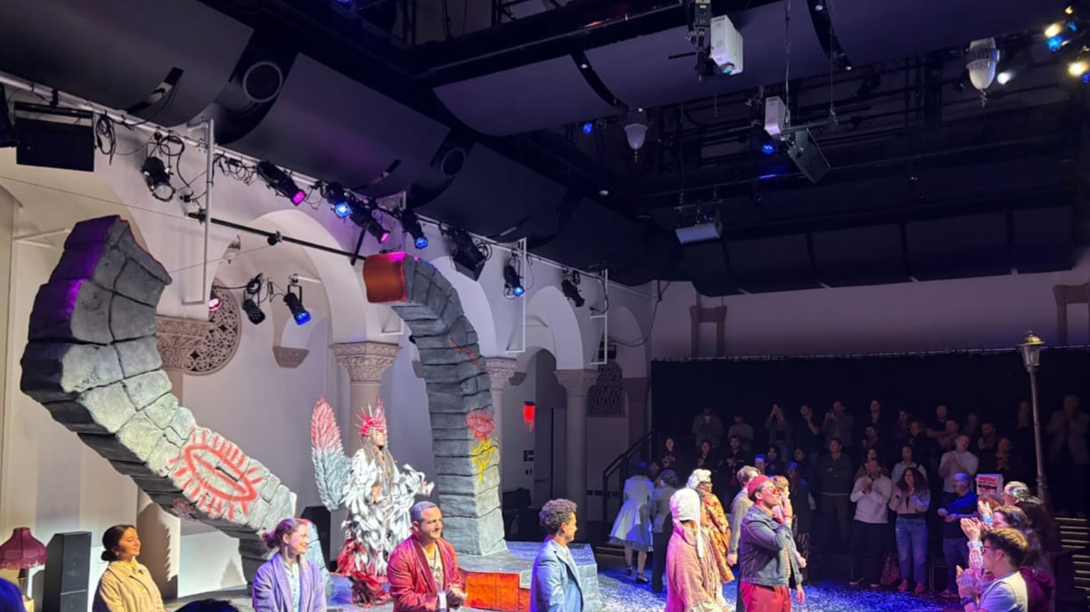 Actors bow on stage while the audience stands and claps. The costumes are colorful with characters in robes.