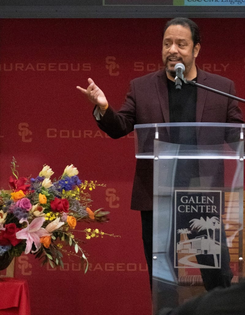 Photo of a man speaking at a podium with a red background behind him.