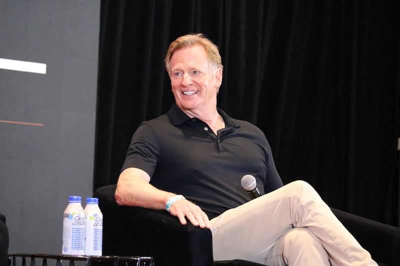 Roger Goodell sits in a black chair with legs crossed and smiles at someone offscreen. He wears a black polo shirt and khaki pants and holds a microphone.