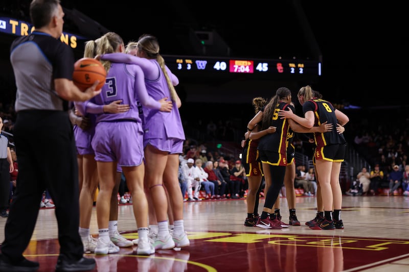 Washington and USC huddle on court. Washington wears bright purple uniforms with black lettering while USC wears black uniforms with cardinal and gold lettering. A referee can also be seen on court holding a basketball near the Washington huddle.