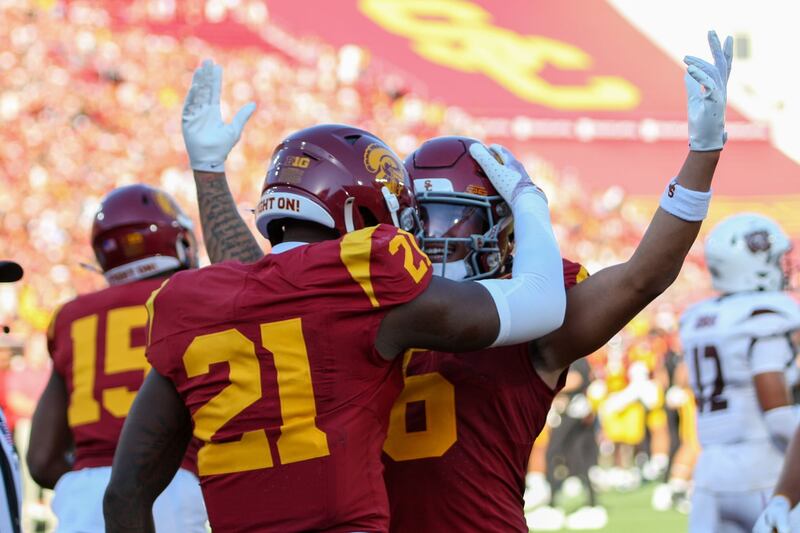 Bryan Jackson (21) celebrates with a teammate; Jackson has his hand on the helmet of his teammate, who is raising both arms in the air as if to celebrate a touchdown. Both wear cardinal USC football jerseys and helmets with gold accents.