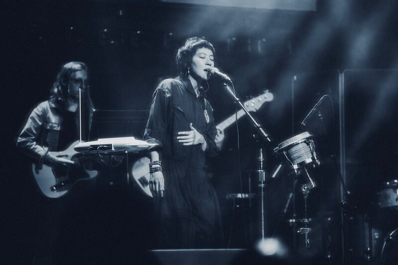 Monochrome photo of a woman singing on stage in front of a pair of guitar players