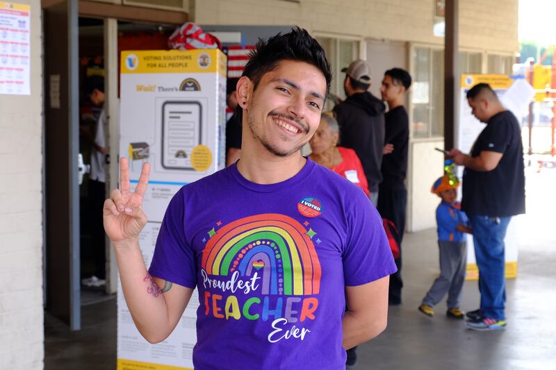 A man in a purple shirt with a rainbow on it smiling and holding up a peace sign.