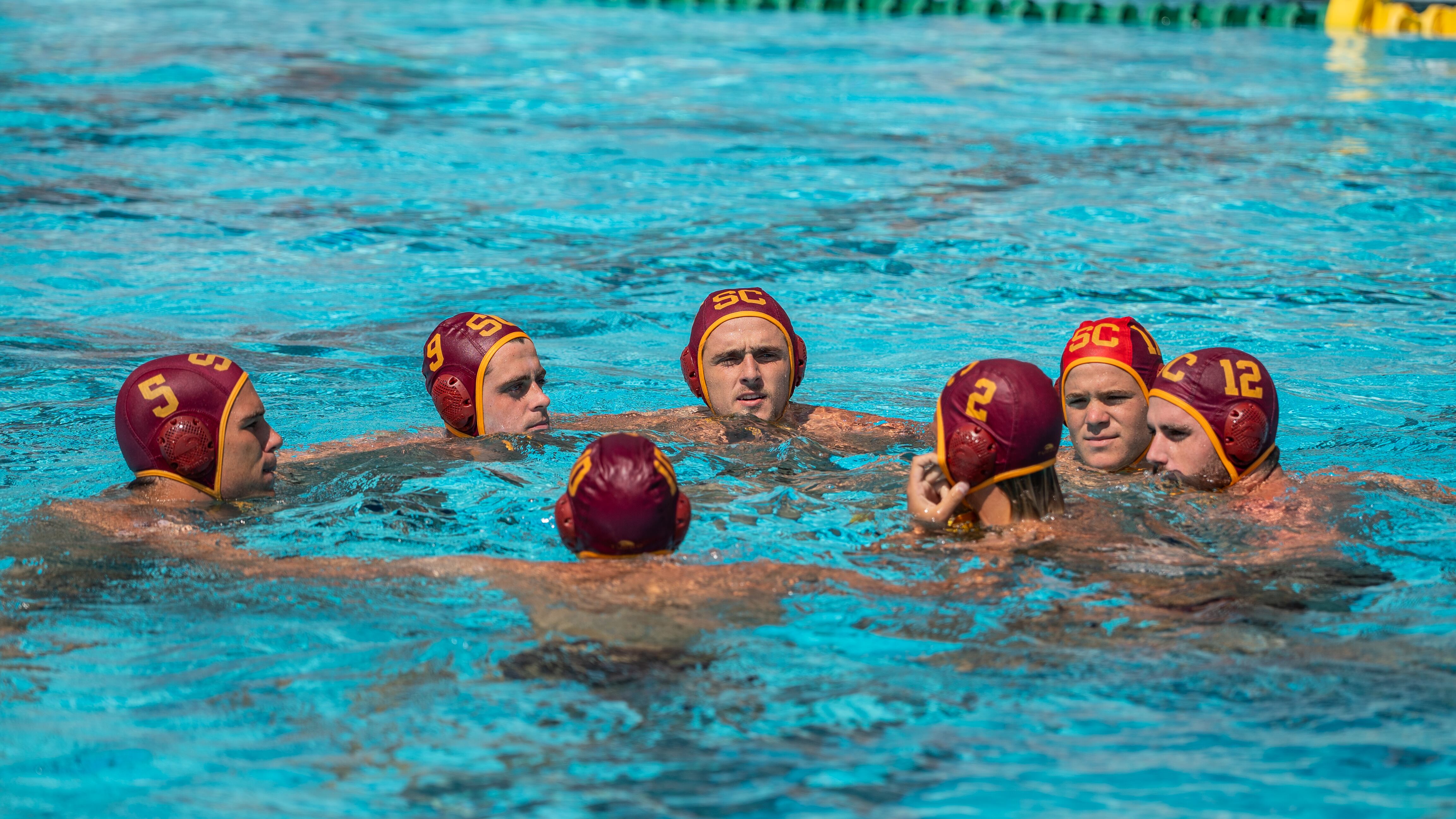 USC men's water polo team huddles in a circle in the water. They wear cardinal and gold swimming caps with their respective number on them and appear to be talking to each other.