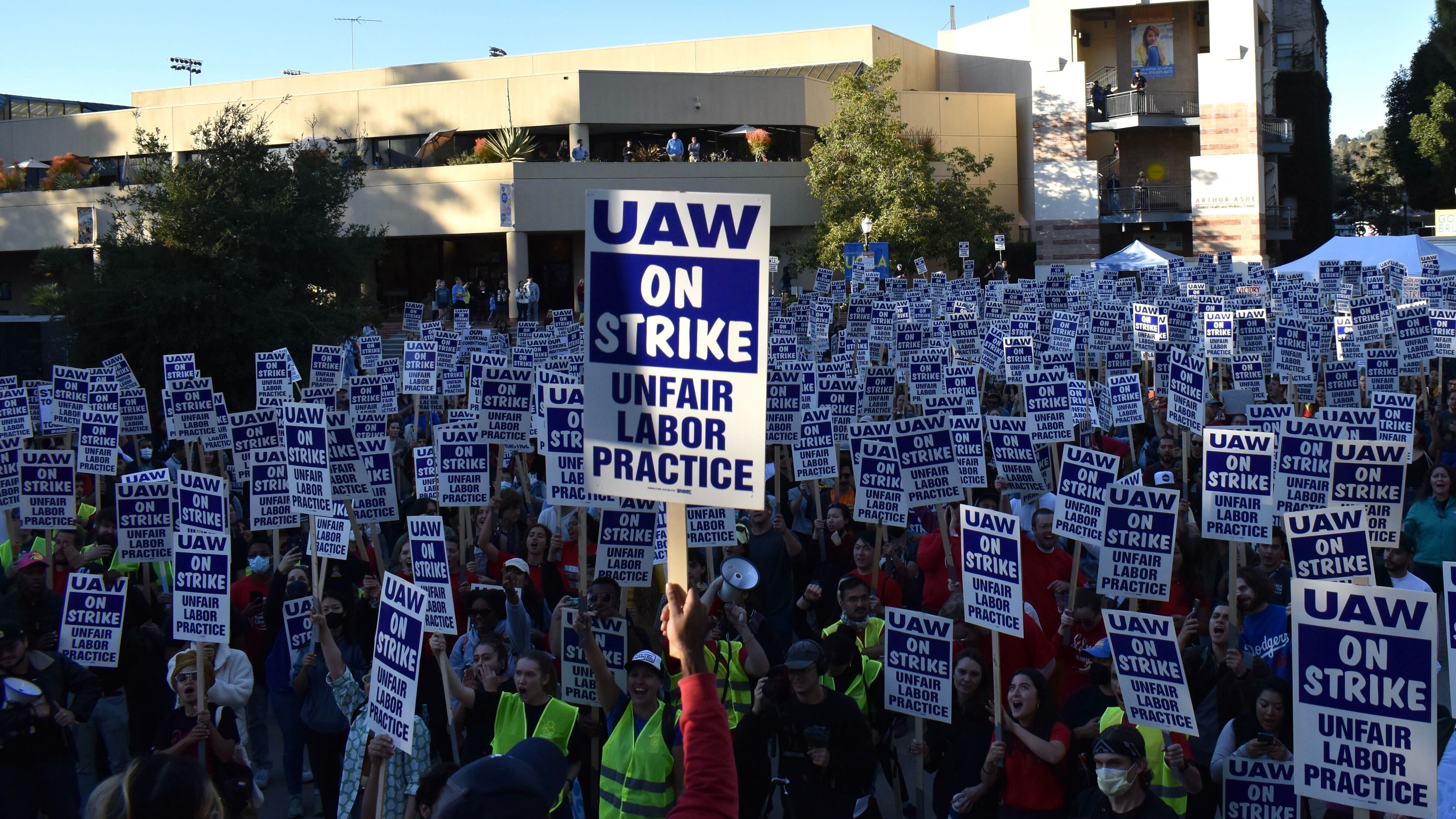 A photo of many striking student workers with white and blue signs that say "UAW on strike, unfair labor practice." One sign stands out from the rest.