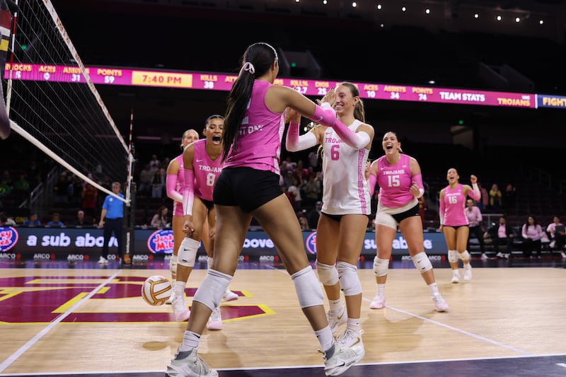 USC women's volleyball players celebrate on Galen Center's court in the foreground; they wear pink and white volleyball jerseys.