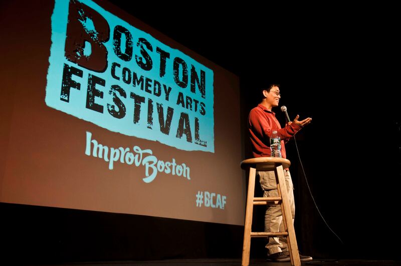 Photo of a man talking on stage by a stool in front of a screen