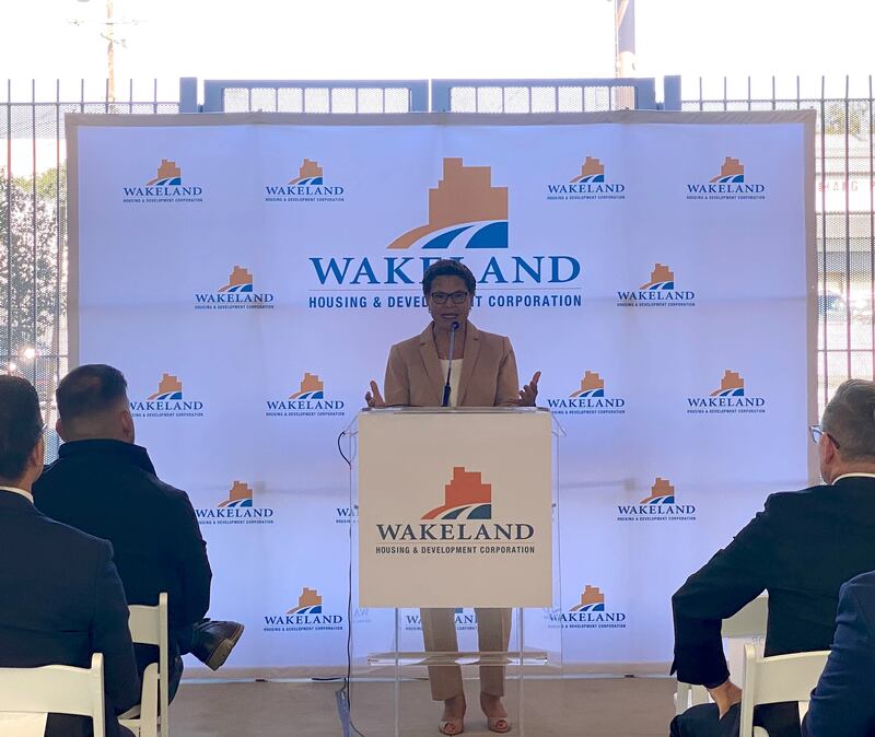 Karen Bass stands behind a lectern and in front of a white backdrop saying "WAKELAND" as she delivers remarks into a microphone to a small crowd of developers and city officials.