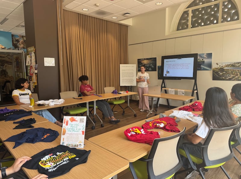 Students sit around desks with shirts in front of them. A volunteer stands in front of a screen showing the students how to repair their clothes.