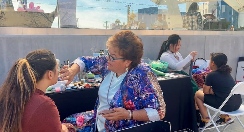 Therese is wearing a purple shawl and a blue shirt. She is applying paint to a girl's face with her fingers. There are other craft booths behind her.