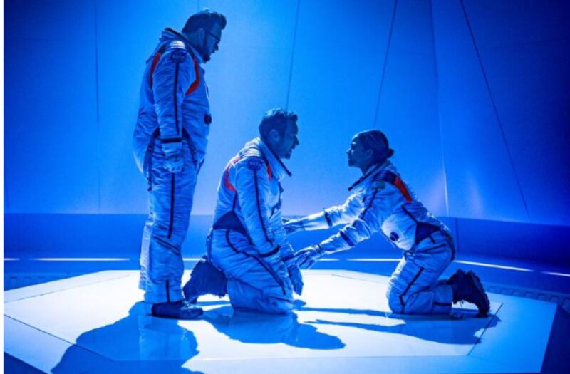 Three astronauts in a blue room. One stands over two who are on the floor on their knees.