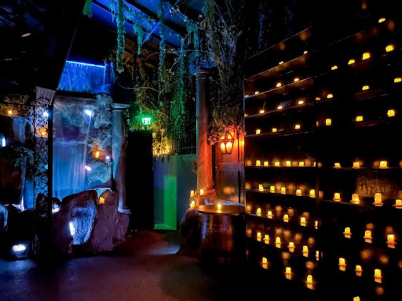 A dark room with fantastical decor, including a waterfall, colorful lighting and a wall of illuminated drinks.