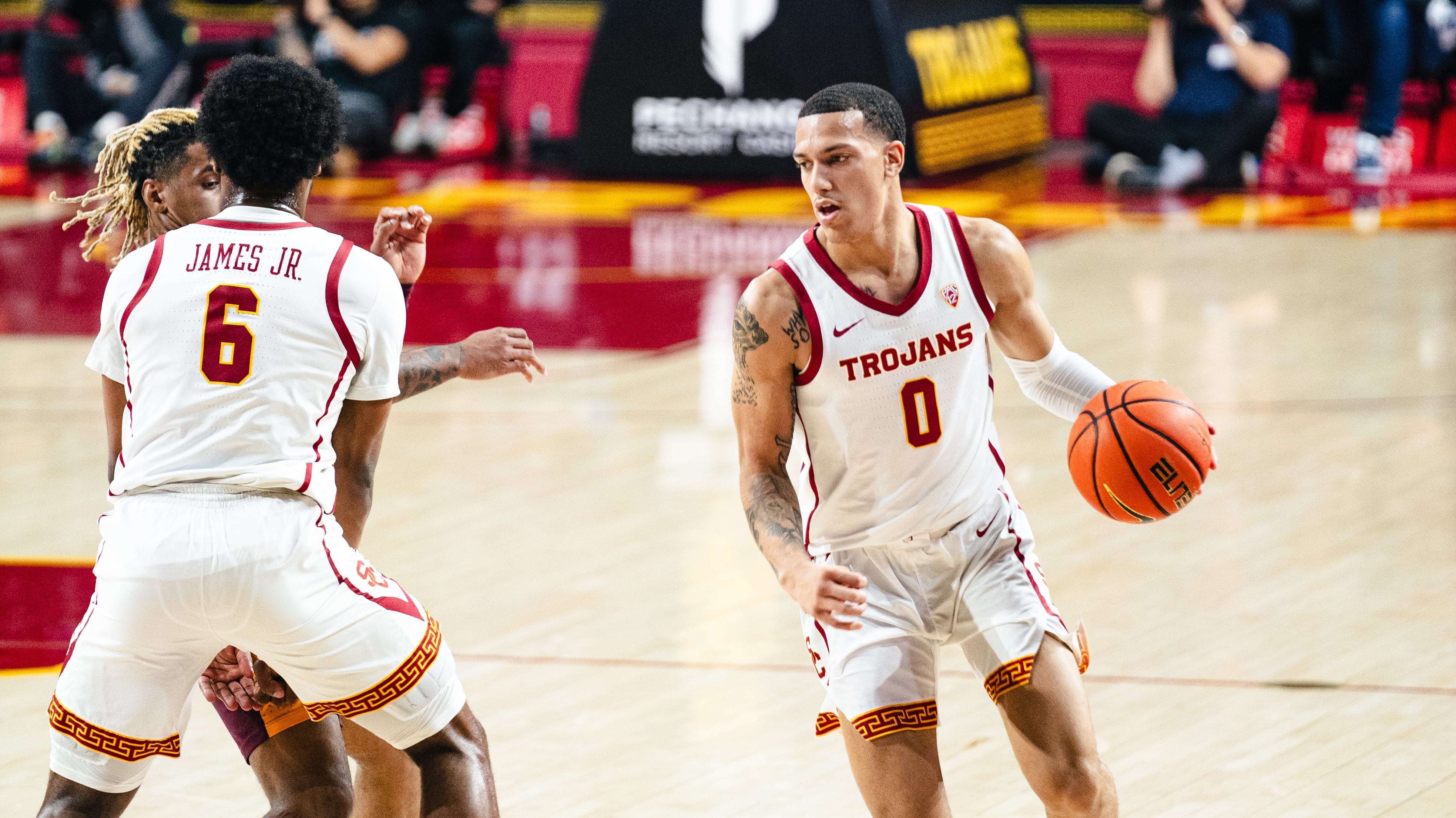 Kobe Johnson, wearing a white Trojans uniform, dribbles the ball with his left hand.
