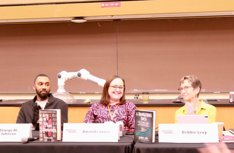 Amanda is smiling and wearing a black and pink shirt. Two panelists to her right and left both smile at her.